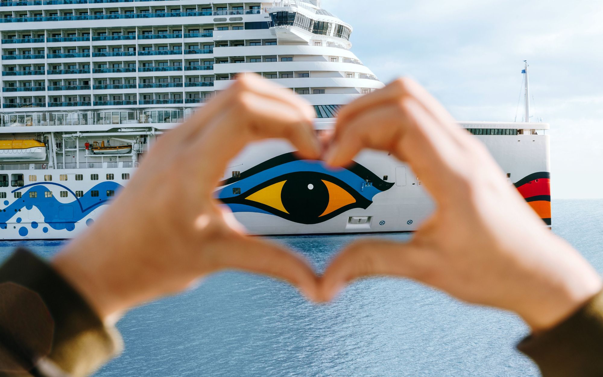 Hände formen ein Herz vor dem AIDA Kussmund-Schiff auf dem Meer